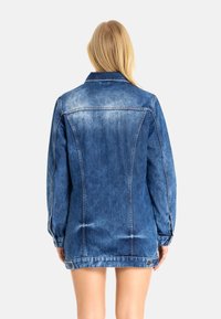 Een spijkerjack in mediumblauw met lange mouwen, een spitse kraag en knoopsluiting. Heeft vervaging en stikdetails door het hele jack.