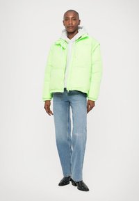 Neon zöld puffermellény elülső cipzárral és zsebekkel, egy szürke kapucnis pulóver fölé rétegezve. A modellen halványkék farmernadrág és fekete magassarkú cipő van.