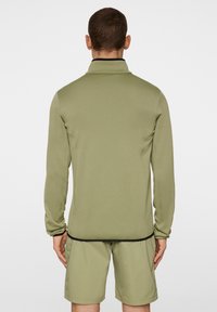 Długi rękaw oliwkowej zielonej bluzki z wysokim kołnierzem, wykonanej z gładkiego materiału, z czarnym wykończeniem na dole i przy mankietach, połączonej z pasującymi szortami.