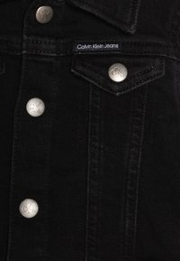 Črna traper jakna z srebrnimi kovinskimi gumbi in CK logotipom. Ima koničast ovratnik in žep na prsih z detajli šivanja.