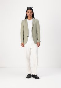 Jachetă verde deschis cu rever clasic, două butoane negre, purtată peste un tricou alb, asortată cu pantaloni crem și pantofi negri.