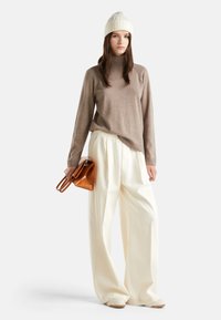 Žena oblečená v béžovém roláku, širokých smetanových kalhotách, bílém pleteném čepici, držící hnědou koženou kabelku, stojící na bílém pozadí.