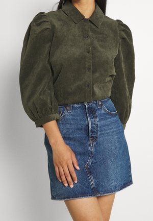 Mujer con una camisa de pana color verde oliva abotonada con mangas abullonadas y una minifalda vaquera azul.