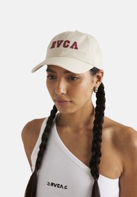 Cappello beige a righe con logo RVCA, dotato di visiera curva e cinturino regolabile. Indossato con una canotta bianca, mostrando un'acconciatura a treccia.