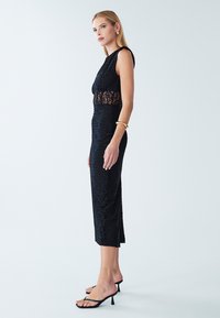 Robe en dentelle noire à la silhouette cintrée, sans manches, avec une partie médiane en dentelle transparente, longueur genou, associée à des sandales à talons hauts noires.