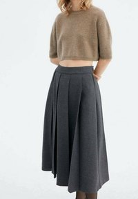 Donna che indossa un maglione corto marrone e una gonna midi plissettata grigia scura a vita alta, su uno sfondo chiaro e neutro.