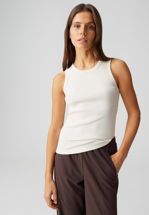 Donna con capelli lunghi castani che indossa una canotta bianca a coste senza maniche e pantaloni marrone scuro, in piedi con una mano in tasca davanti a uno sfondo grigio.