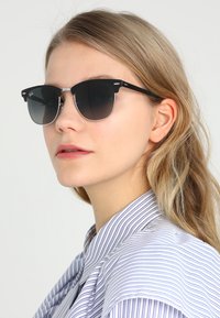 Ochelari de soare negri cu lentile gri degradé, punte metalică și brațe din plastic. Purtați de o persoană cu păr lung și ondulat, îmbrăcată cu un tricou cu dungi.
