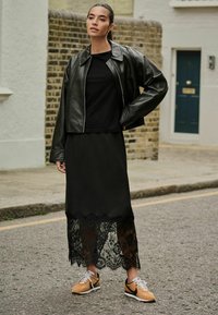 Black leather cropped jacket, black lace-trimmed midi skirt, and beige sneakers with black accents. Smooth textures, layered design.