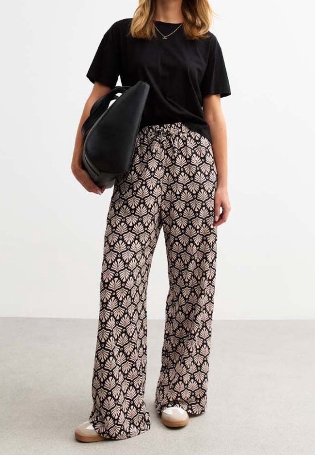 Femme portant un t-shirt noir et un pantalon large à motifs, tenant un grand sac à main noir, debout sur un sol et un fond unis.