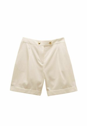  Béžové krátke nohavice s vysokým pásom, ktorý je upravený na mieru, s ohnutými lemami, prednými záhybmi, bočnými vreckami a dvoma zlatými gombíkmi na opasku.