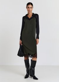 Vestito verde a sottoveste con bordi in pizzo, indossato sopra una camicia nera a maniche lunghe. Stivali neri al ginocchio e una borsa abbinata completano il look.