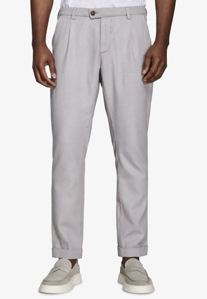 Mężczyzna ubrany w jasnoszare, dopasowane spodnie z podwiniętymi nogawkami i beżowe mokasyny zamszowe, stojący na jednolitym tle.