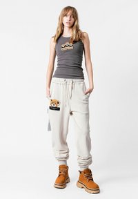 Camiseta sin mangas gris con un diseño bordado, combinada con pantalones de chándal beige que tienen un parche de oso de peluche y un bolsillo negro. Botas color tostado con suelas gruesas.