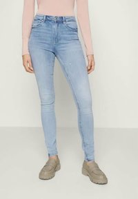 Jeans skinny azuis claros feitos de denim, com um clássico design de cinco bolsos e um ligeiro desgaste. Usados com sapatos slip-on bege.