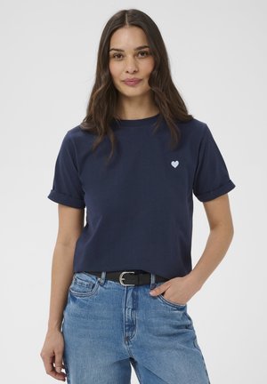 Femme aux longs cheveux bruns portant un t-shirt bleu marine avec un petit emblème en forme de cœur et un jean bleu clair avec une ceinture noire, la main dans la poche.