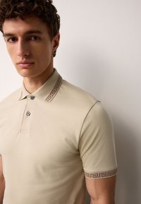 Beiges Poloshirt mit einem dekorativen braunen griechischen Schlüsselmuster am Kragen und an den Ärmelsäumen, ausgestattet mit zwei Knöpfen und einer glatten Textur.