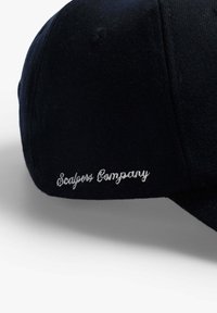 Zwarte pet van zacht stof, met een gebogen rand en witte geborduurde tekst "Scalpers Company" aan de zijkant.