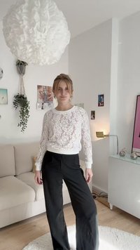 Λευκή δαντελένια μπλούζα με μακριά μανίκια και στρογγυλή λαιμόκοψη, συνδυασμένη με μαύρα παντελόνια σε φαρδιά γραμμή, τοποθετημένα σε μια μινιμαλιστική εσωτερική διακόσμηση.