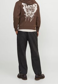 Felpa con cappuccio marrone e grafica di una farfalla bianca sul retro, abbinata a pantaloni larghi scuri e stivali marroni stringati.
