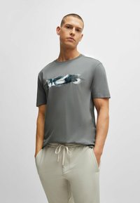 Homme portant un T-shirt graphique gris et un pantalon beige à cordon de serrage, les mains dans les poches, se tenant devant un fond gris clair uniforme.