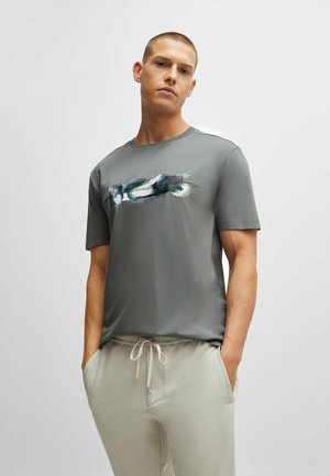 Homme portant un T-shirt graphique gris et un pantalon beige à cordon de serrage, les mains dans les poches, se tenant devant un fond gris clair uniforme.