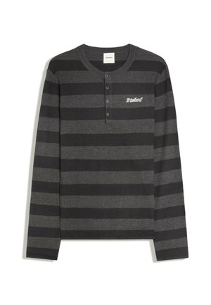 Uomo dalla pelle scura che indossa una camicia nera a righe a maniche lunghe, cintura con borchie, pantaloni scuri, occhiali da sole, che tiene il manico di una borsa su sfondo chiaro e uniforme.