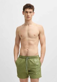 Men's olive green swim shorts with an elastic waistband, drawstring, side pockets, and a large logo printed on the left leg.