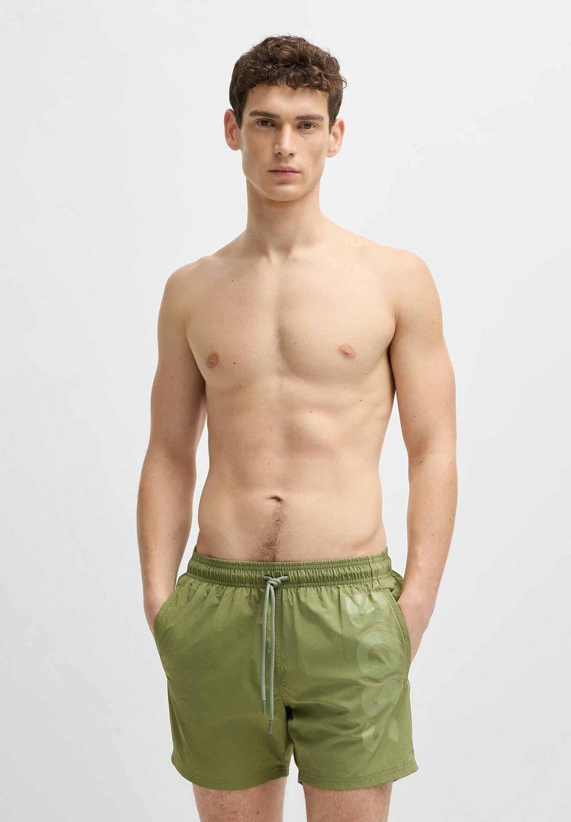 Men's olive green swim shorts with an elastic waistband, drawstring, side pockets, and a large logo printed on the left leg.