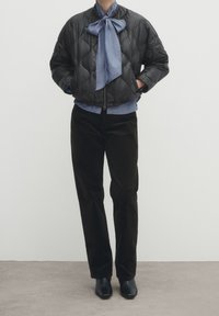 Person iført en sort quilted jakke, blå bluse med en stor sløjfe, sorte bukser og sorte hælede ankelstøvler, der står på et gråt gulv.