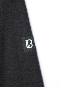 Fekete bolyhos anyag, texturált felülettel, zöld címkével a hónalon, fehér betűkkel "B" felirattal.