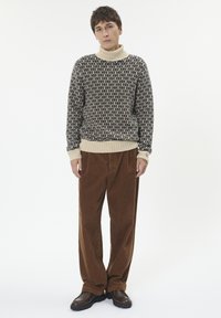 Mladic stoji, odjeven u bež pulover s uzorkom i visokim ovratnikom, labave smeđe baršunaste hlače i tamnosmeđe cipele, na bijeloj pozadini.