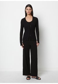 Woman standing against a white wall wearing a black ribbed cardigan, black wide-leg pants, and black open-toe slip-on sandals.