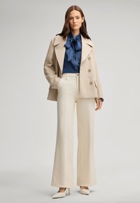Cappotto beige doppiopetto, blusa blu scuro con cravatta, pantaloni larghi color crema e décolleté a punta color crema. Tessuto morbido, design strutturato.