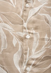 Tissu marron clair avec des motifs de feuilles botaniques blanches et trois boutons blancs le long de la couture centrale.