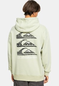 Sudadera verde clara con un gran diseño gráfico negro en la parte trasera que presenta olas y montañas. Fabricada en un tejido suave con capucha ajustable.