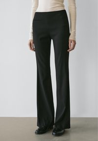 Woman wearing black high-waisted straight-leg trousers, beige long-sleeve top, and black shoes standing on a gray floor against a white wall.