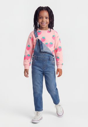 Smiling girl with braided hair wears pink strawberry sweatshirt, denim overalls with one strap undone, and white high-top sneakers, standing.