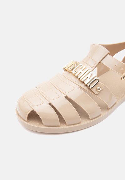 Sandale en gelée beige brillante avec plusieurs brides horizontales et détail du logo MOSCHINO doré sur la bride supérieure.