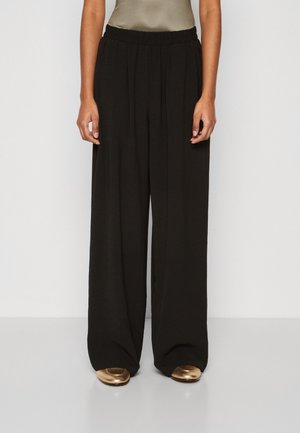 Personne portant un pantalon large noir et des chaussures plates métalliques dorées, debout sur un fond blanc uni.