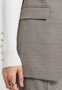 Hnědá a bílá látka saka s kostkovaným vzorem s žebrovaným bílým rukávem, zdobená zlatými knoflíky a detailem boční kapsy.
