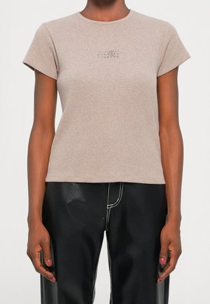 Persoon die een beige kortemouwen top draagt met kleine gedrukt cijfers op de borst en een zwarte broek met witte stiksels.