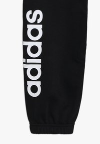 Zwarte sportieve joggers van gladde stof, met een opvallend wit "adidas" logo verticaal langs het linkerbeen en elastische boorden.