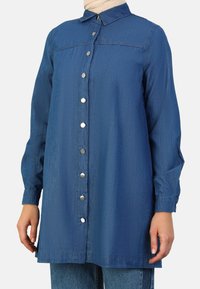 Camisa de mezclilla en azul oscuro, con cierre frontal de botones plateados, mangas largas y un cuello clásico. Textura suave y corte relajado.