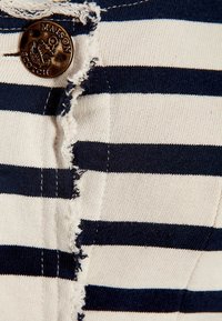 Tejido a rayas en azul marino y crema con un borde deshilachado, que presenta un botón redondo oscuro con un logotipo de ancla y detalles de costura visibles.