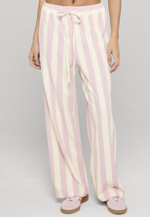 Donna che indossa pantaloni larghi a righe verticali rosa e bianche con coulisse in vita e sneakers rosa, in piedi davanti a uno sfondo semplice.