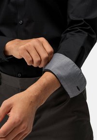 Chemise noire avec des poignets à motif pied-de-poule gris contrastant. Le tissu a une texture lisse et la chemise se ferme avec des boutons.
