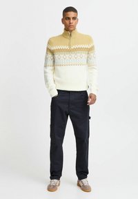 Light yellow and white patterned knit sweater with a high collar, paired with black cargo pants and brown sneakers with white laces.