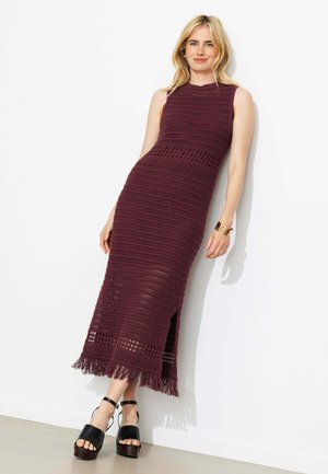 Femme appuyée contre un mur, portant une robe maxi en crochet bordeaux sans manches avec un ourlet à franges et des sandales compensées noires.