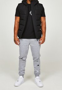 Svart vadderad väst med huva, svart grafisk t-shirt, grå sweatpants med färgfläckar och vita sneakers. Avslappnad, atletisk klädstil.